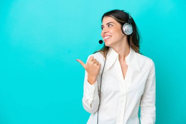Telemarketer Italian woman working with a headset isolated on blue background pointing to the side to present a product