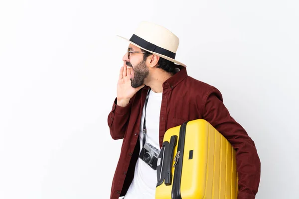 Traveler man man with beard holding a suitcase over isolated white background shouting with mouth wide open to the lateral