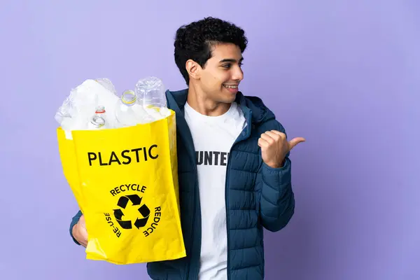 Young Venezuelan man holding a bag full of plastic bottles pointing to the side to present a product