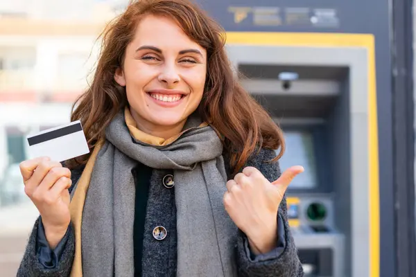 Brunette woman holding a credit card at outdoors pointing to the side to present a product