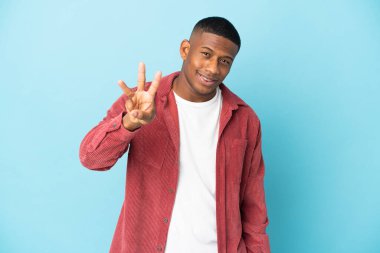 Mavi arka planda izole edilmiş genç Latin adam mutlu ve parmaklarıyla üçe kadar sayıyor.