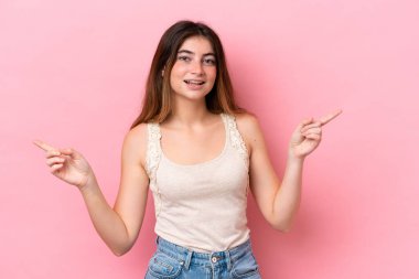 Pembe arka planda izole edilmiş genç beyaz kadın parmak uçlarını işaret ediyor ve mutlu.
