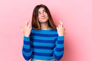 Pembe arka planda izole edilmiş genç beyaz kadın parmaklarını çapraz yapıp en iyisini diliyor.