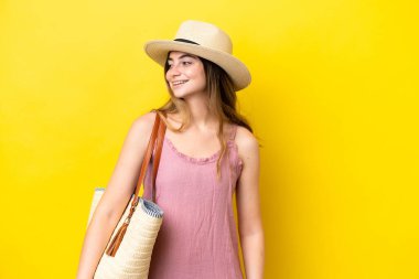 Sarı arka planda izole edilmiş bir plaj çantası tutan genç beyaz kadın yana bakıyor ve gülümsüyor.