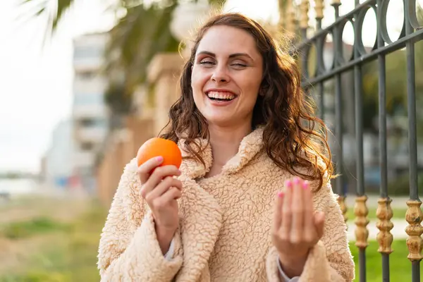 Young caucasian woman holding an orange at outdoors inviting to come with hand. Happy that you came