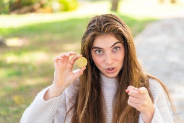 Açık havada Bitcoin tutan güzel beyaz bir kadın şaşırmış ve ön tarafı işaret ediyor.