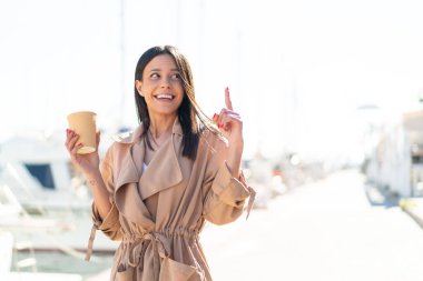 Açık havada elinde kahve tutan genç bir kadın çözümün farkına varmak için parmağını kaldırıyor.