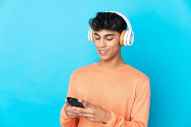 Genç adam izole edilmiş mavi arka planda müzik dinliyor ve cep telefonu arıyor.