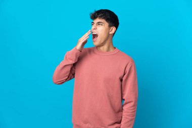 Genç adam izole edilmiş mavi arka planda esniyor ve elleriyle ağzını kapatıyor.