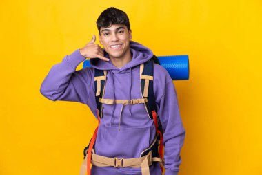 İzole edilmiş sarı arka planda büyük bir sırt çantası olan genç dağcı telefon hareketinde bulundu. Bana geri dönüş işareti ver.