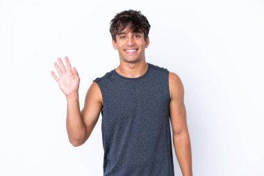 Beyaz arka planda izole edilmiş beyaz tenli genç bir adam mutlu bir ifadeyle selamlıyor.