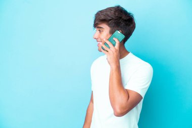 Kafkasyalı genç adam mavi arka planda izole edildi. Cep telefonuyla biriyle sohbet ediyor.