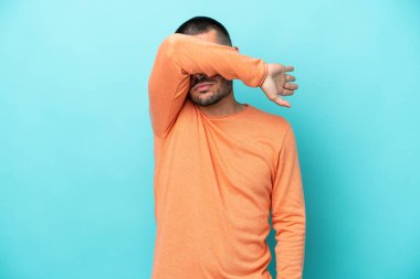 Genç beyaz adam mavi arka planda izole edildi. Gözlerini elleriyle kapadı.