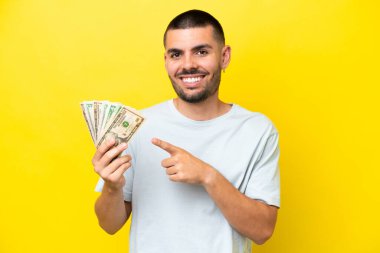 Young caucasian man taking a lot of money isolated on yellow background pointing to the side to present a product