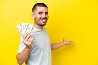 Kafkasyalı genç adam, sarı arka planda izole edilmiş bir sürü para alıyor. Davet etmek için ellerini yana uzatıyor.