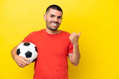 Futbol oynayan beyaz bir genç sarı arka planda izole edilmiş bir şekilde bir ürün sunmak için yan tarafa bakıyor.