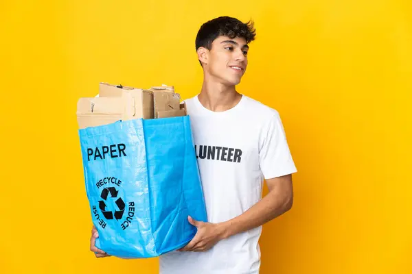 Man holding a recycling bag full of paper to recycle over isolated yellow background looking side