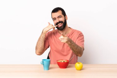 Kafkasyalı bir adam masada kahvaltı yapıyor telefon jesti yapıyor ve ön tarafı işaret ediyor..