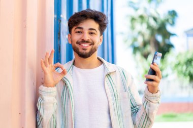 Yakışıklı bir Arap açık havada cep telefonu kullanıyor. Tamam işareti parmaklarıyla gösteriyor.