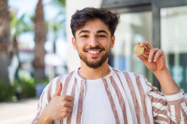 Yakışıklı bir Arap açık havada Bitcoin tutuyor. Başparmakları havada çünkü iyi bir şey oldu.