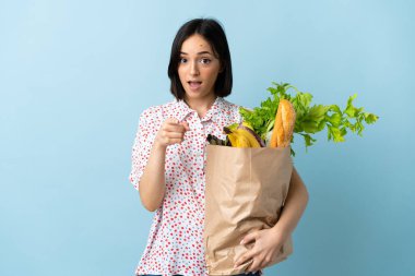 Elinde alışveriş çantası tutan genç bir kadın şaşırmış ve ön tarafı işaret ediyor.