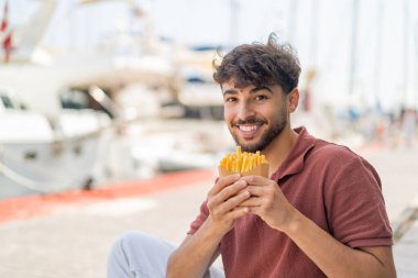 Genç Arap yakışıklı adam açık havada neşeli bir ifadeyle kızarmış cips yiyor.