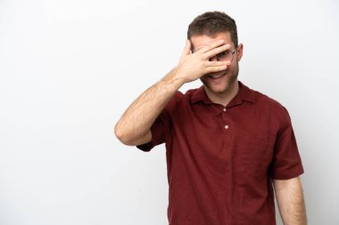 Beyaz arka planda izole edilmiş beyaz tenli genç bir adam. Gözleri elleriyle kaplıyor ve gülümsüyor.