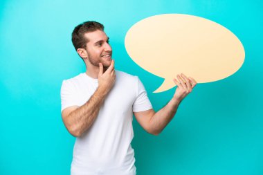 Young caucasian man isolated on blue background holding an empty speech bubble and pointing it