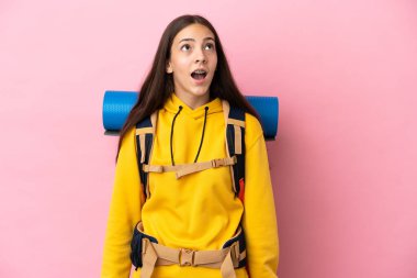 Pembe arka planda izole edilmiş büyük sırt çantalı dağcı kız yukarı bakıyor ve şaşırmış bir ifadeyle