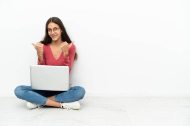 Genç Fransız kız dizüstü bilgisayarıyla yerde oturuyor. Başparmakları havada ve gülümsüyor.