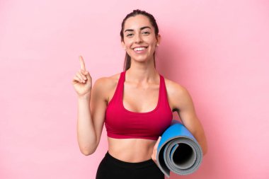 Genç beyaz kadın yoga derslerine gidiyor. Elinde pembe arka planda izole edilmiş bir minder var. En iyinin işareti olarak parmağını kaldırıyor.
