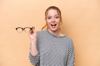 Young caucasian girl with glasses isolated on beige background with surprise and shocked facial expression