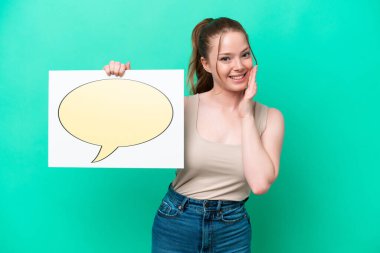 Genç beyaz kadın yeşil arka planda izole edilmiş. Elinde konuşma balonu ikonu olan bir levha var ve bağırıyor.