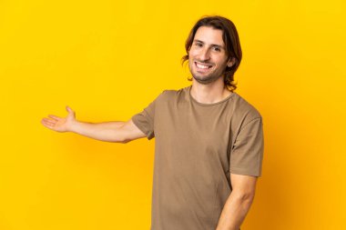Sarı arka planda izole edilmiş genç ve yakışıklı bir adam davet edildiği için ellerini yana uzatıyor.