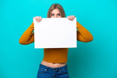 Young caucasian woman isolated on blue background holding an empty placard and hiding behind it