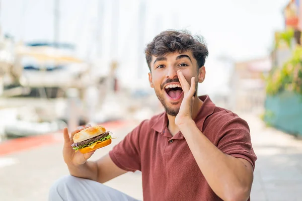 若いアラビアのハンサムな男は 開いた口で叫んで屋外でハンバーガーを保持 — ストック写真