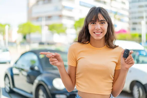 Young woman holding car keys at outdoors surprised and pointing finger to the side