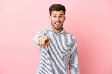 Pembe arka planda izole edilmiş beyaz, yakışıklı genç adam şaşırmış ve ön tarafı işaret ediyor.