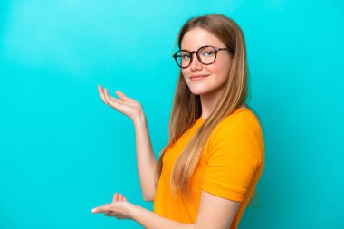Mavi arka planda izole edilmiş genç beyaz kadın, davet edilmek için ellerini yana uzatıyor.