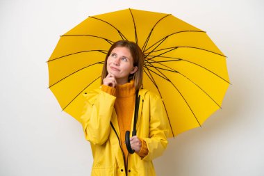 Yağmura dayanıklı ceketi ve şemsiyesi olan genç bir İngiliz kadın beyaz arka planda izole edilmiş ve yukarı bakıyor.