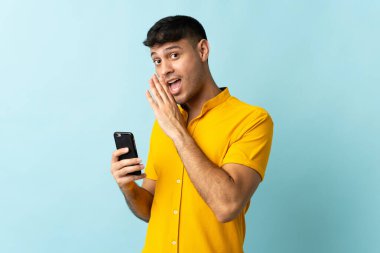 Cep telefonu kullanan genç bir Kolombiyalı mavi arka planda bir şeyler fısıldıyor.
