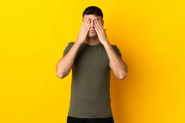Sarı arka planda izole edilmiş genç Kolombiyalı adam gözlerini elleriyle kapatıyor.