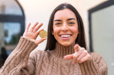 Açık havada Bitcoin tutan genç bir kadın kendinden emin bir ifadeyle seni işaret ediyor.