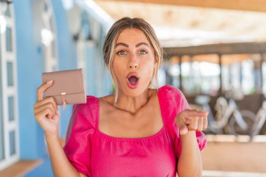 Açık havada cüzdan tutan genç sarışın kadın şaşırmış ve ön tarafı işaret ediyor.