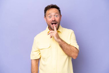 Mor arka planda izole edilmiş orta yaşlı beyaz bir adam sessizlik işareti gösteriyor. Parmağını ağzına sokuyor.