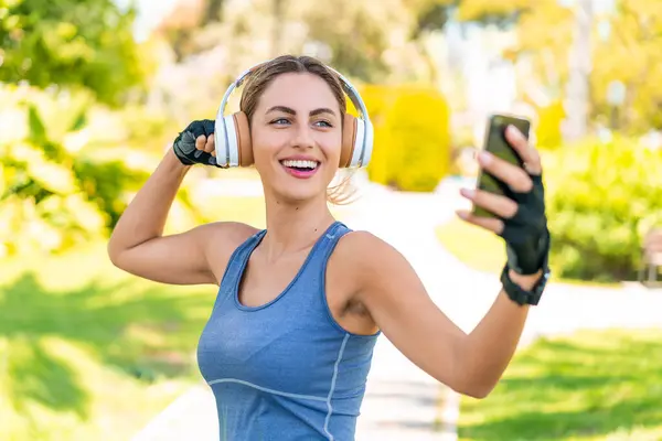 Açık havada spor giysileri giyen ve cep telefonuyla selfie çeken genç sarışın kadın.