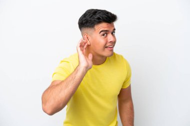 Beyaz arka planda izole edilmiş beyaz tenli yakışıklı bir adam elini kulağına koyarak bir şeyler dinliyor.