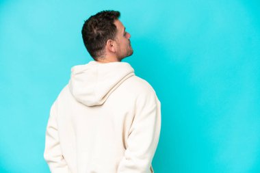 Kafkasyalı yakışıklı genç adam mavi arka planda izole edilmiş ve arkasına bakıyor.