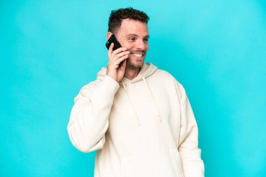 Mavi arka planda izole edilmiş genç beyaz adam cep telefonuyla sohbet ediyor.