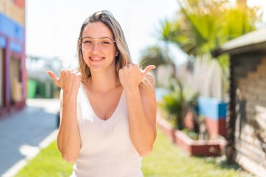 Açık havada genç ve güzel bir kadın. Baş parmakları havada ve gülümsüyor.